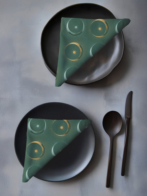 Four-piece cloth napkin set featuring a celestial print of radiant golden crescent moons and pale sunburst orbs against a rich emerald green background. Each napkin displays symmetrical, glowing motifs inspired by lunar phases and cosmic geometry. The fabric is neatly folded to showcase the repeating pattern, with a soft matte finish and fine detailing, perfect for mystical or magical table settings, altar decor, or celestial-inspired dining aesthetics.